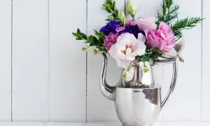 Bukiet różowych i fioletowych eustoma wielkokwiatowa w srebrnym dzbanku na tle białej, drewnianej ściany.