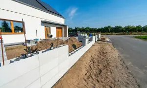 Budowa ogrodzenia z bloczków betonowych, widoczna część domu i plac budowy.