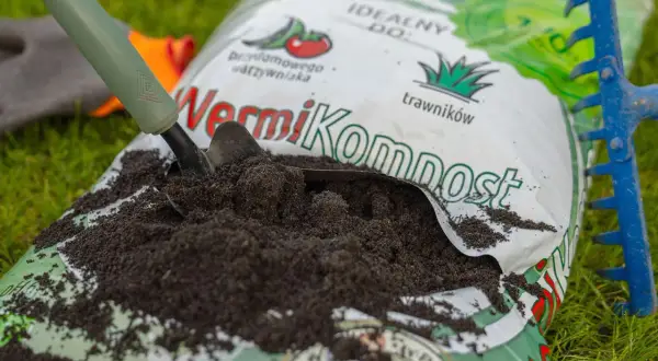 Łopata nabiera wermikompost z otwartej torby na trawie, obok niebieskich grabi.