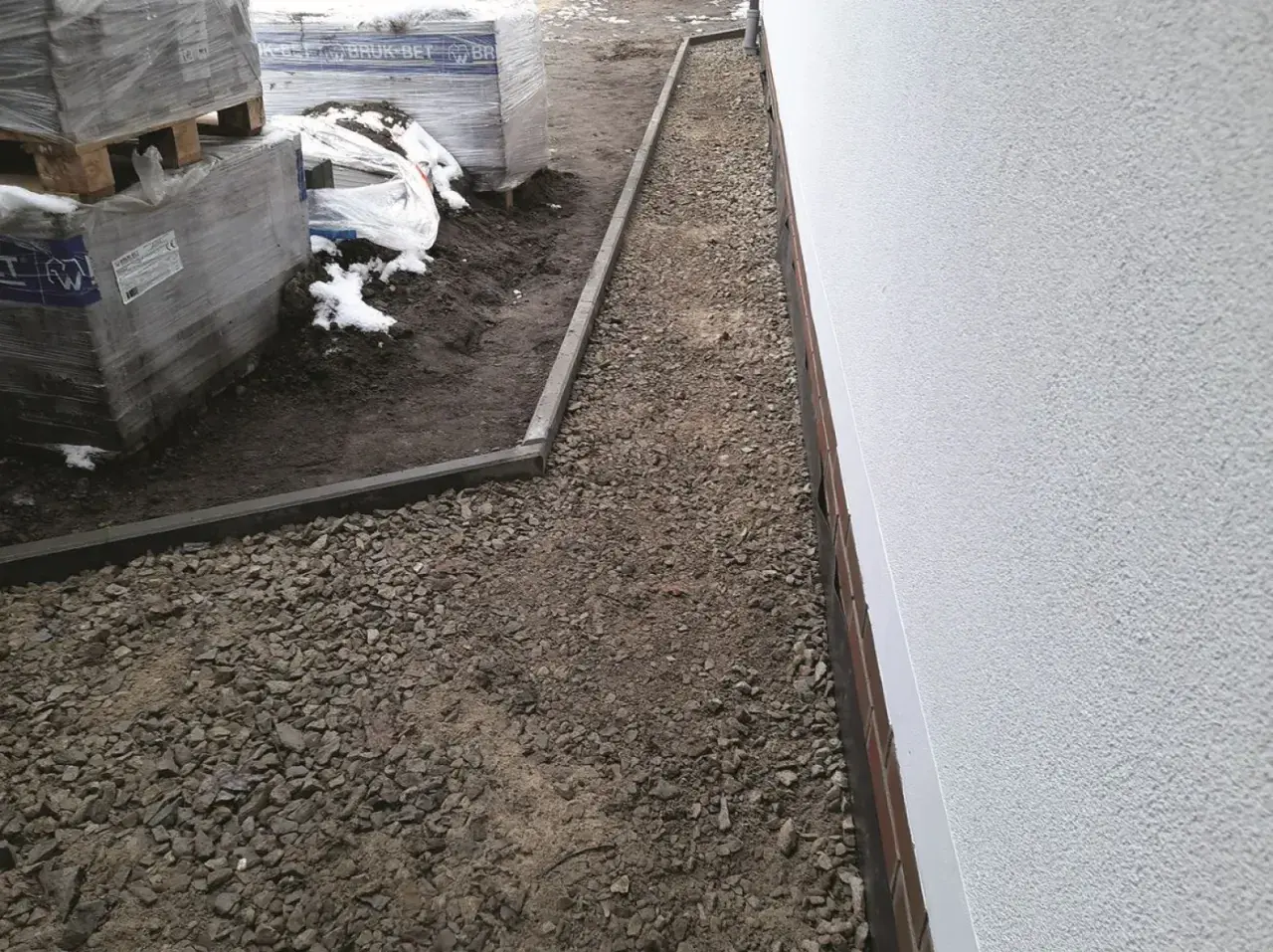 Nawierzchnia mineralna wokół budynku, obok palet z materiałami budowlanymi.