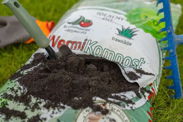 Łopata nabiera wermikompost z otwartej torby na trawie, obok niebieskich grabi.