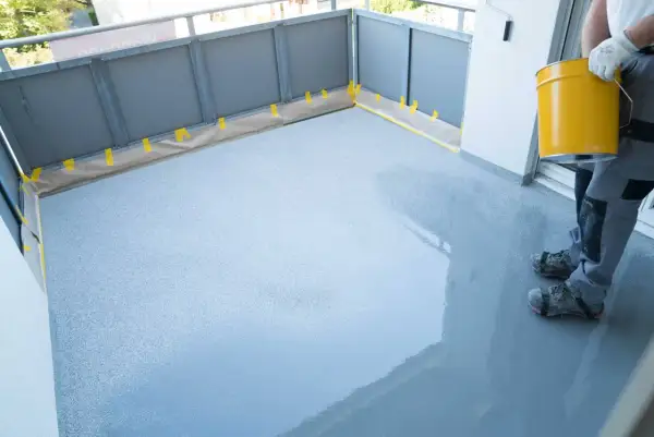 Osoba w rękawicach i specjalnych butach nakłada szarą masę hydroizolacyjną na balkon.