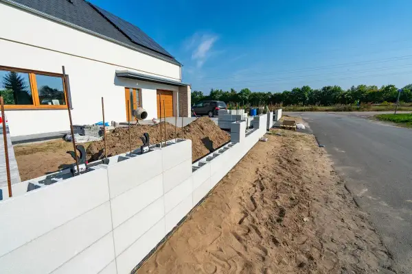 Budowa ogrodzenia z bloczków betonowych, widoczna część domu i plac budowy.